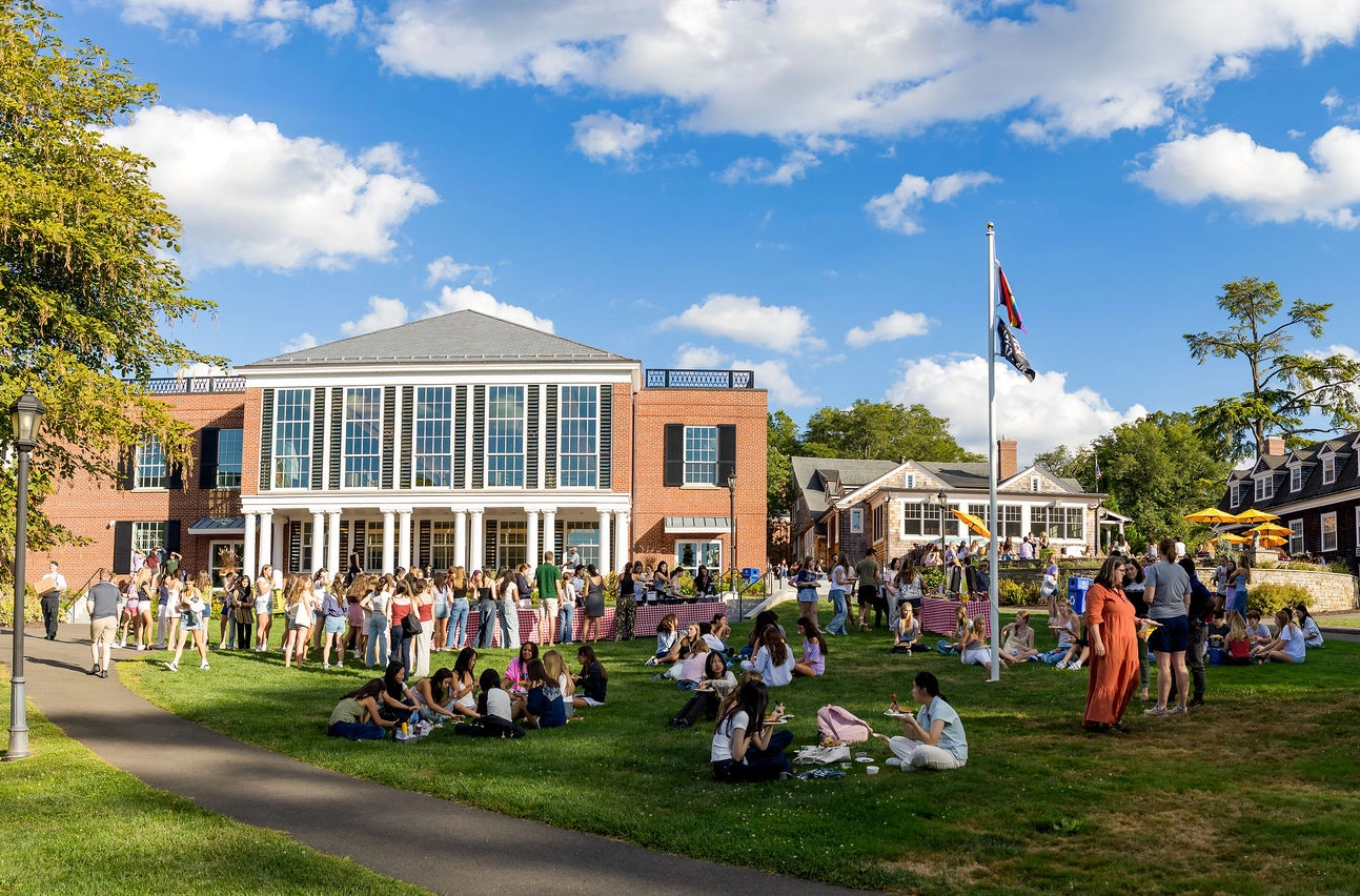 Students gather on a grassy lawn in front of a brick school building under a sunny sky, sitting, talking, and standing in groups, with a flagpole visible nearby.