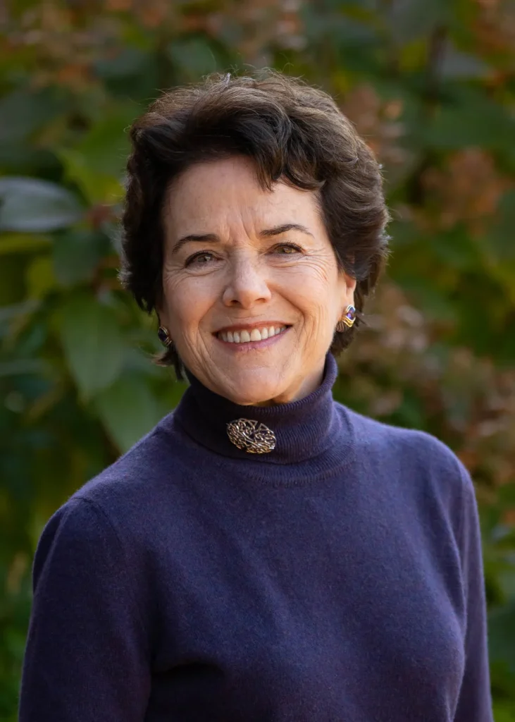 A woman with short dark hair and a purple turtleneck sweater smiles outdoors, standing in front of green foliage.