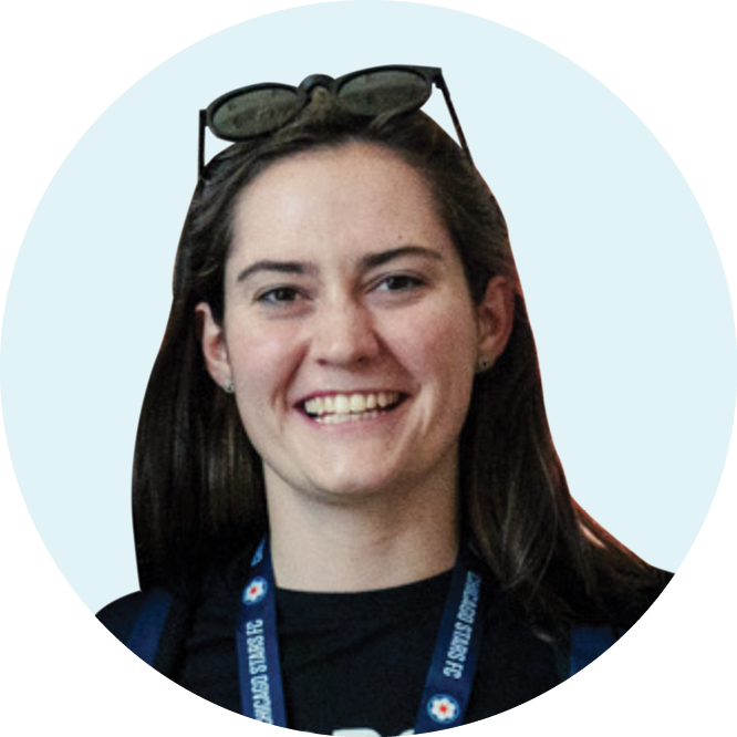 A woman with long brown hair smiles, wearing sunglasses on her head and a lanyard around her neck, against a light background.