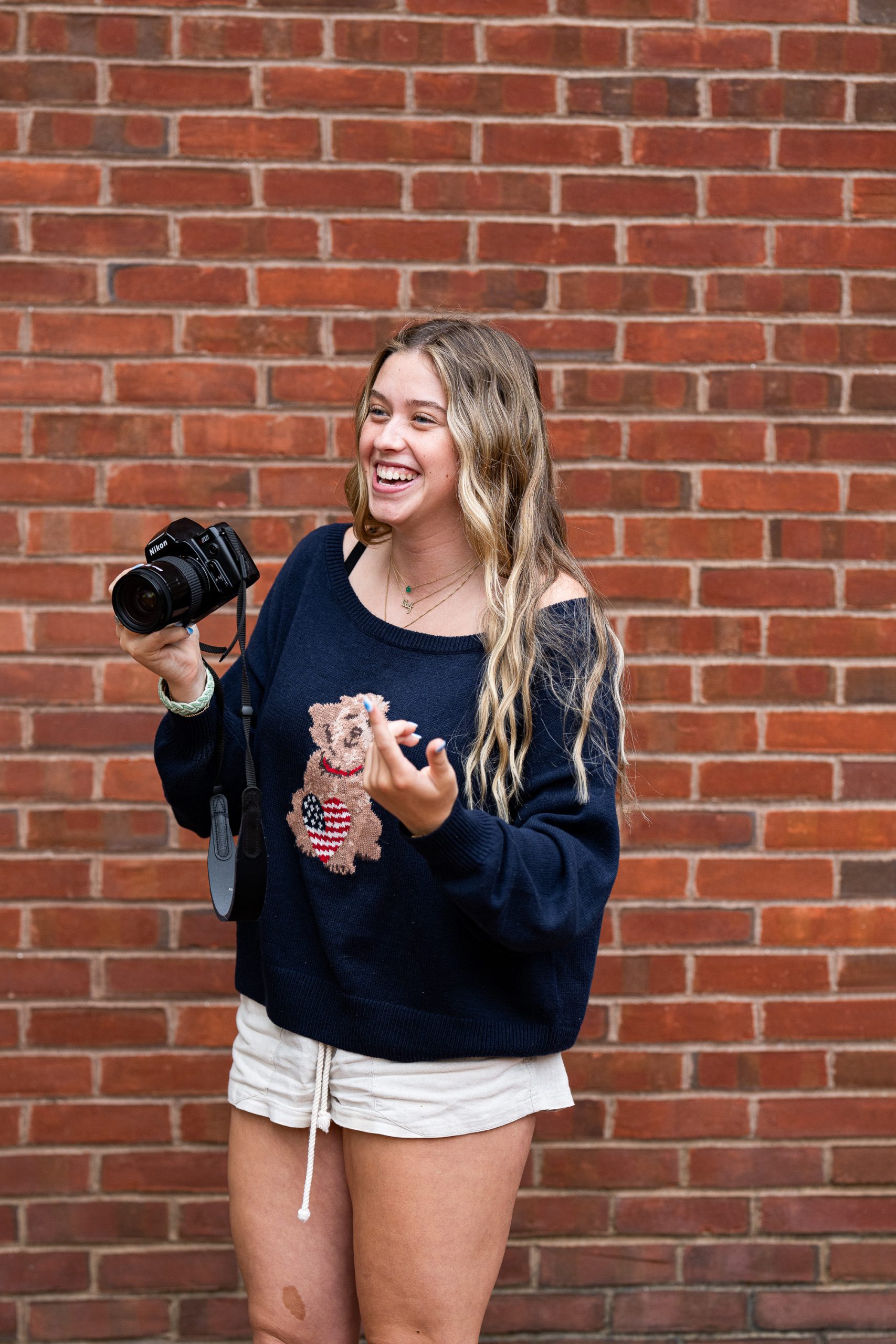 A smiling woman with long hair holds a camera and gestures with her hand, standing in front of a brick wall. She wears a navy sweater with a bear design and light shorts.