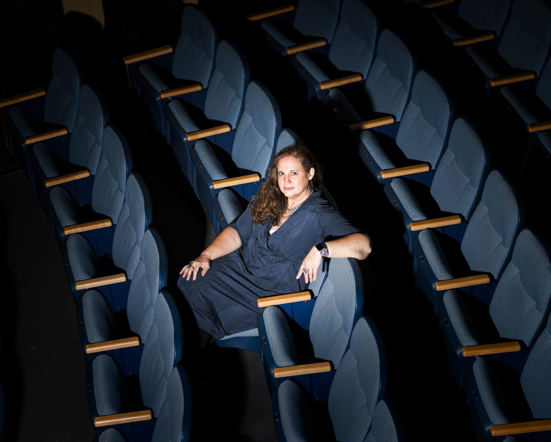 A person with long hair sits alone in a dimly lit auditorium, surrounded by empty blue seats.