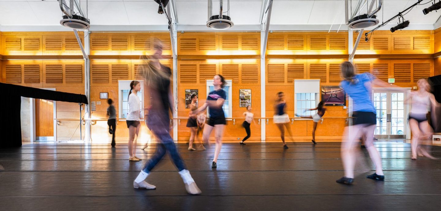 A group of people practice dance in a brightly lit studio with wooden walls. Some figures appear blurred, suggesting movement and energetic choreography.
