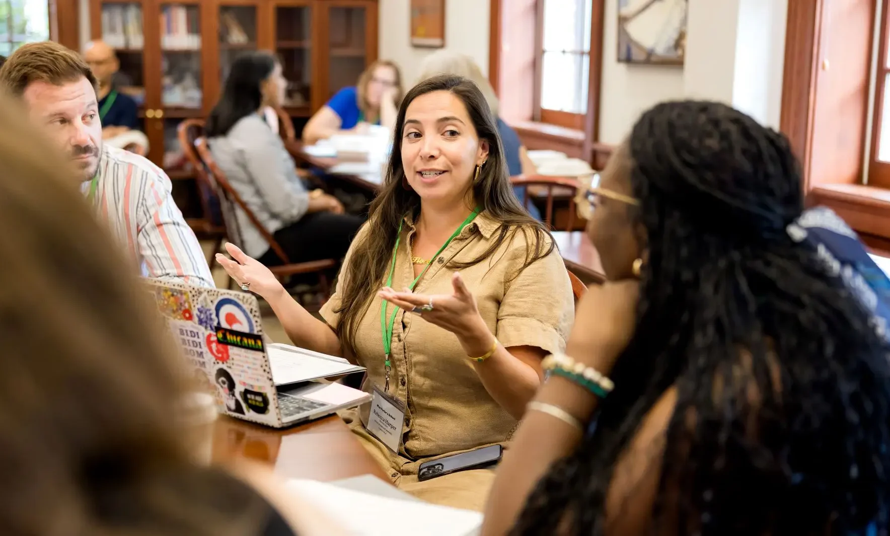 A woman gestures while speaking to a group around a table in a bright room with large windows, bookshelves, and several people engaged in conversation about opportunities for educators.