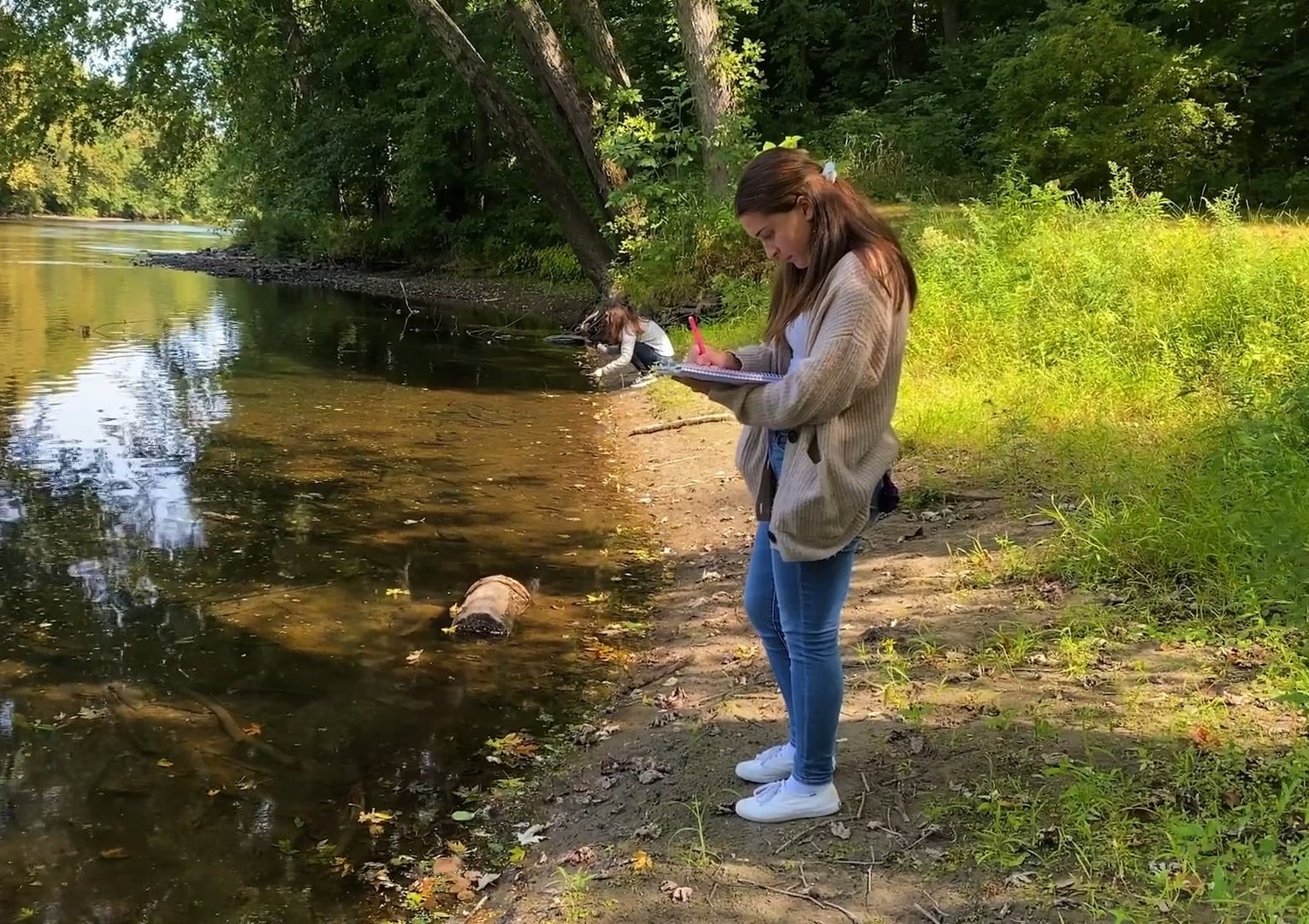 A woman in a beige sweater stands by a calm, tree-lined riverbank, writing or drawing in a notebook. A small dog is near the waters edge. Sunlight filters through the trees, creating a peaceful outdoor scene.