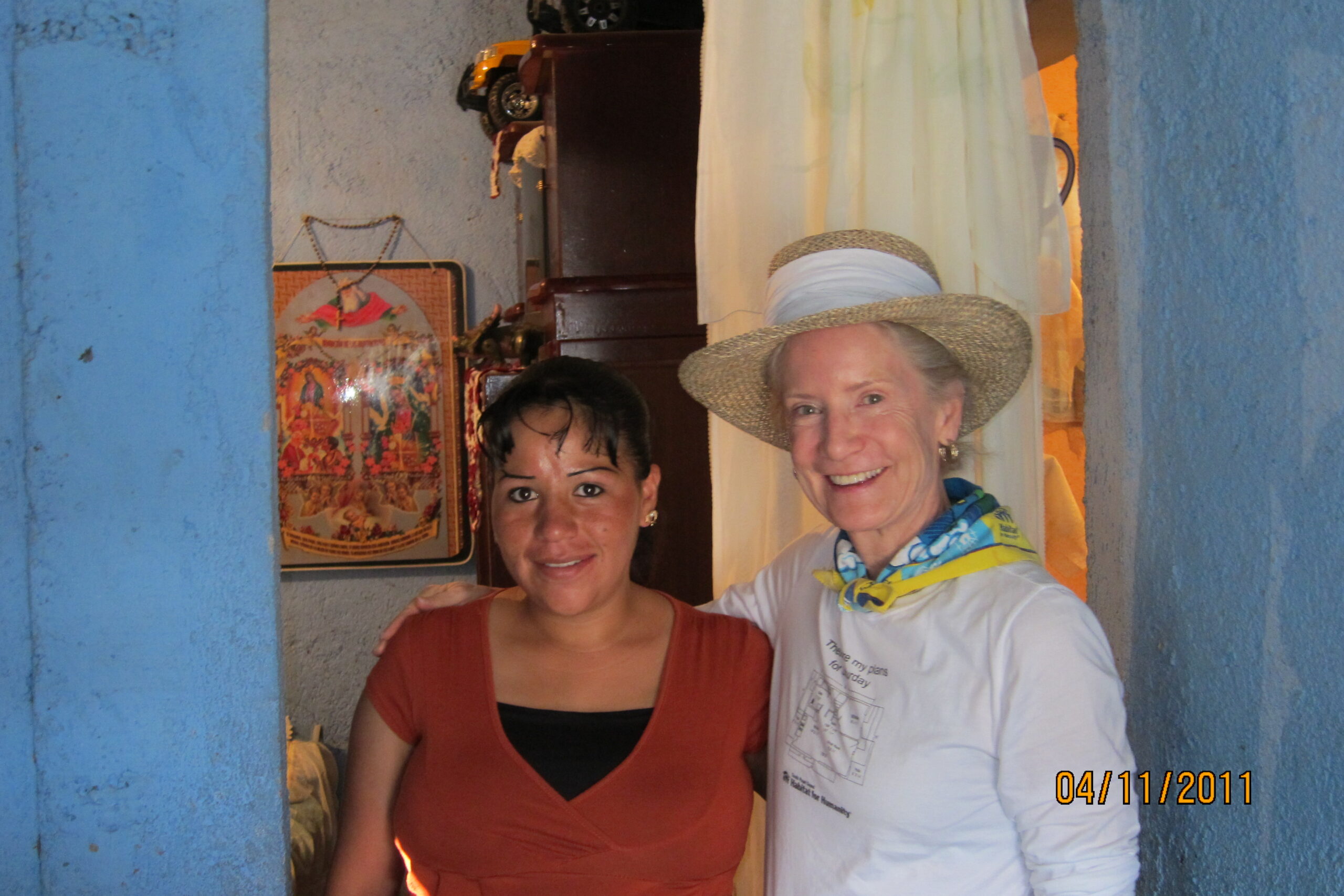 Two women smiling and standing close together indoors, one wearing a straw hat and white shirt, the other in a red top. A vintage sewing machine and a colorful framed picture are visible in the background. Dated 04/11/2011.