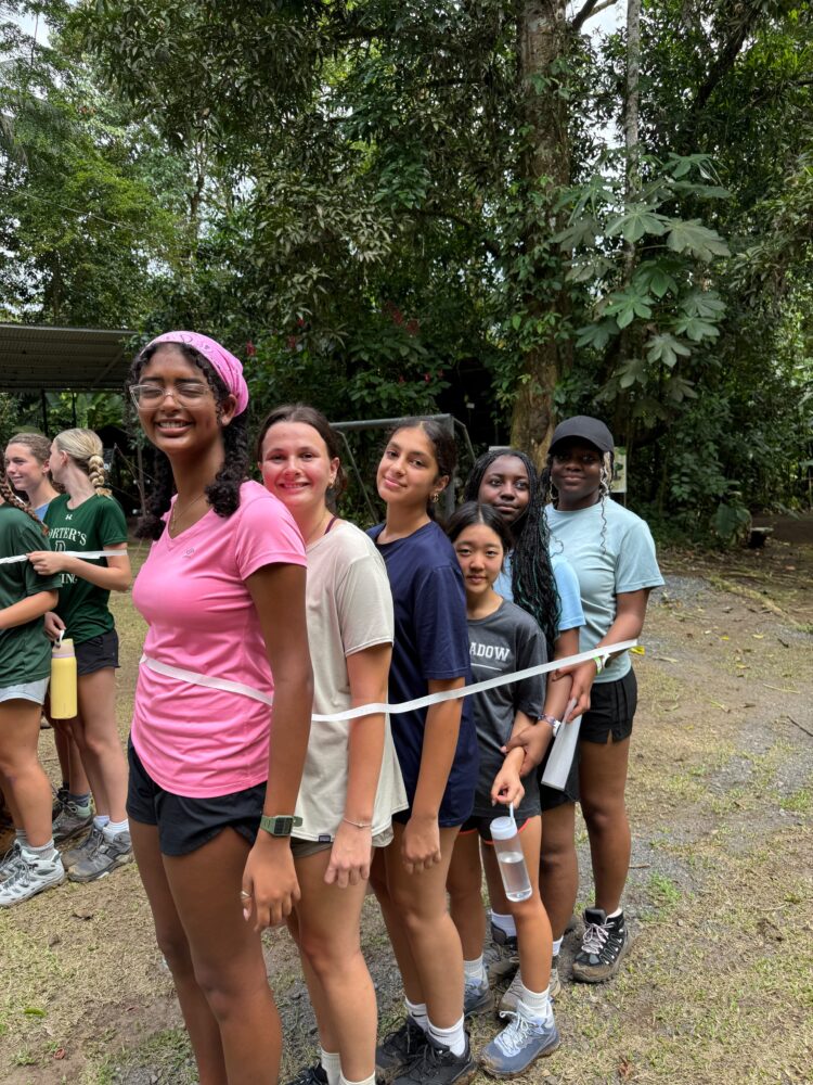 A group of seven young people standing in a line outdoors, connected by a piece of white tape held against their waists.