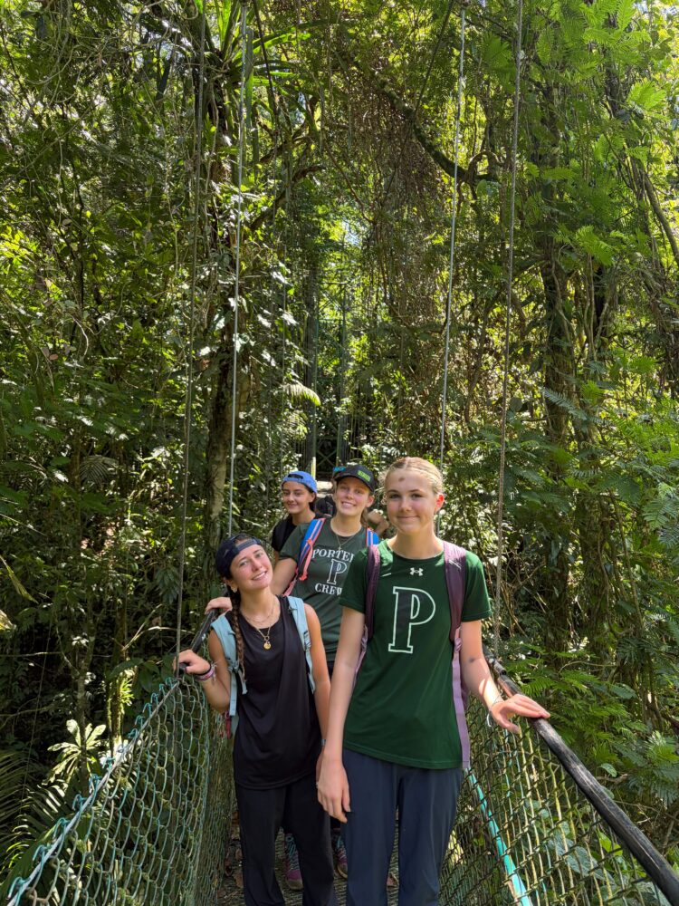 Four young women walk across a suspension bridge in a dense forest.