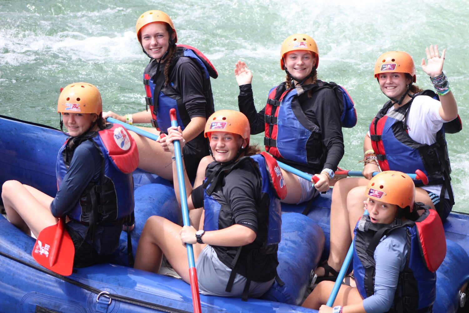 Five people in life vests and helmets sit in a blue raft on whitewater, smiling and waving at the camera.