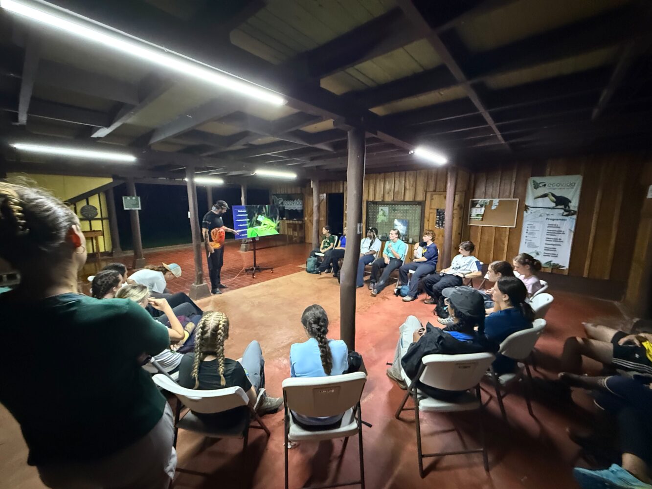 A group of people sit on folding chairs in a wooden lodge, watching a person give a presentation on a monitor.
