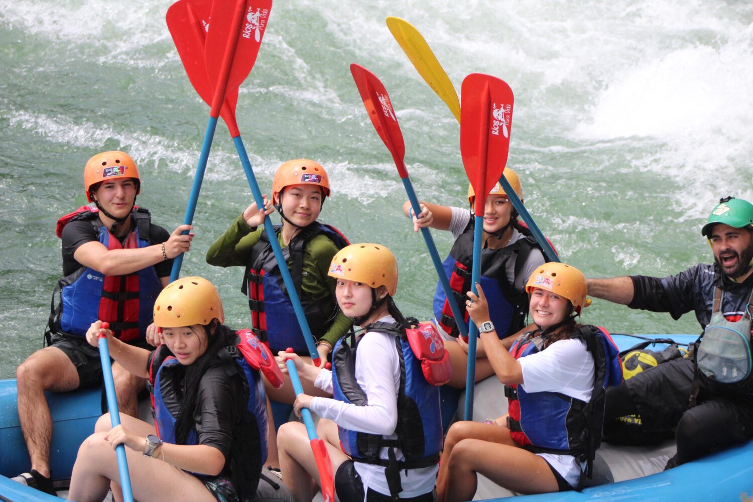 A group of six people in helmets and life jackets holding paddles while sitting in a raft on rushing water.