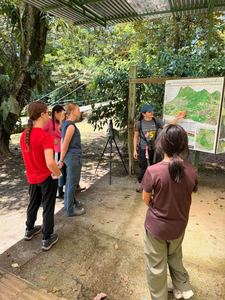 A guide points to a land use map on a wooden stand while speaking to a group of four people in a wooded area.