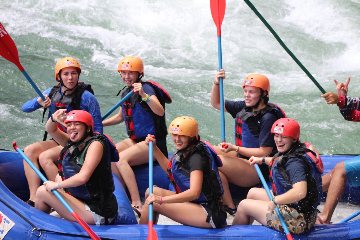 Five people in helmets and life vests paddling a blue raft through whitewater rapids.