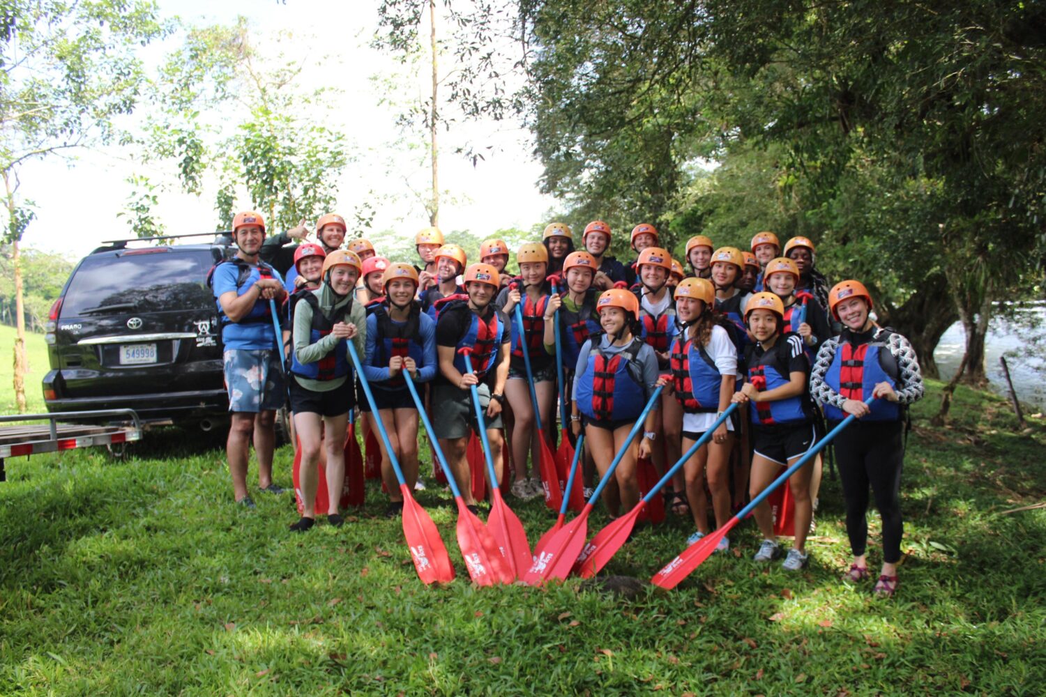 A large group of people in life jackets and helmets pose in a grassy area, holding red rafting paddles in front of them, with a black SUV parked behind them.