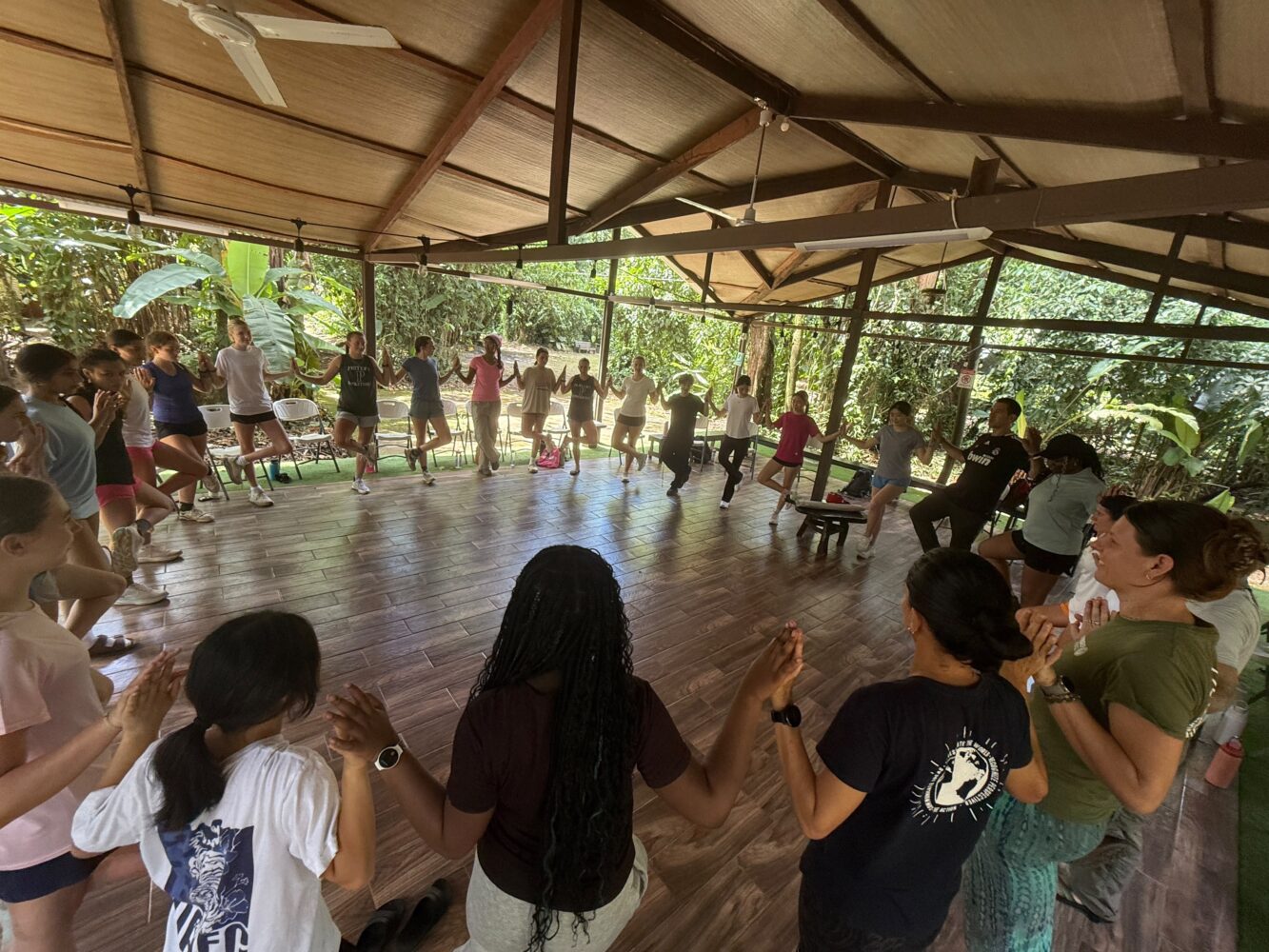 A group of women stand in a large circle in an open-air pavilion, holding hands and facing each other.