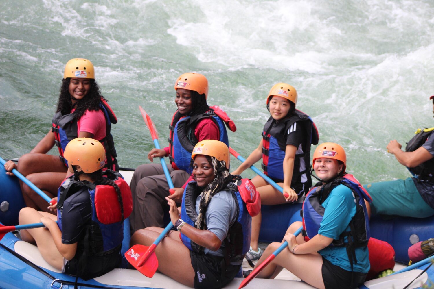 Five people in life jackets and helmets paddle a white water raft through river rapids.