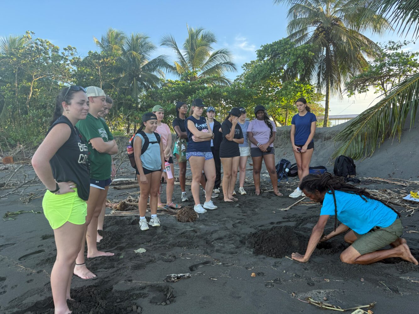 A person kneels on a dark sand beach, digging into the sand while a group of young people stands nearby watching.
