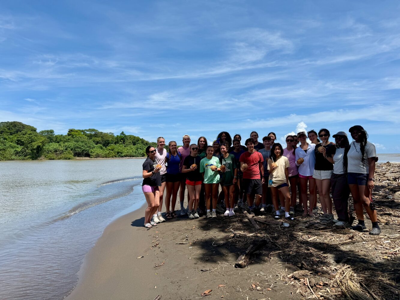 A large group of people standing together on a sandy beach holding coconuts.