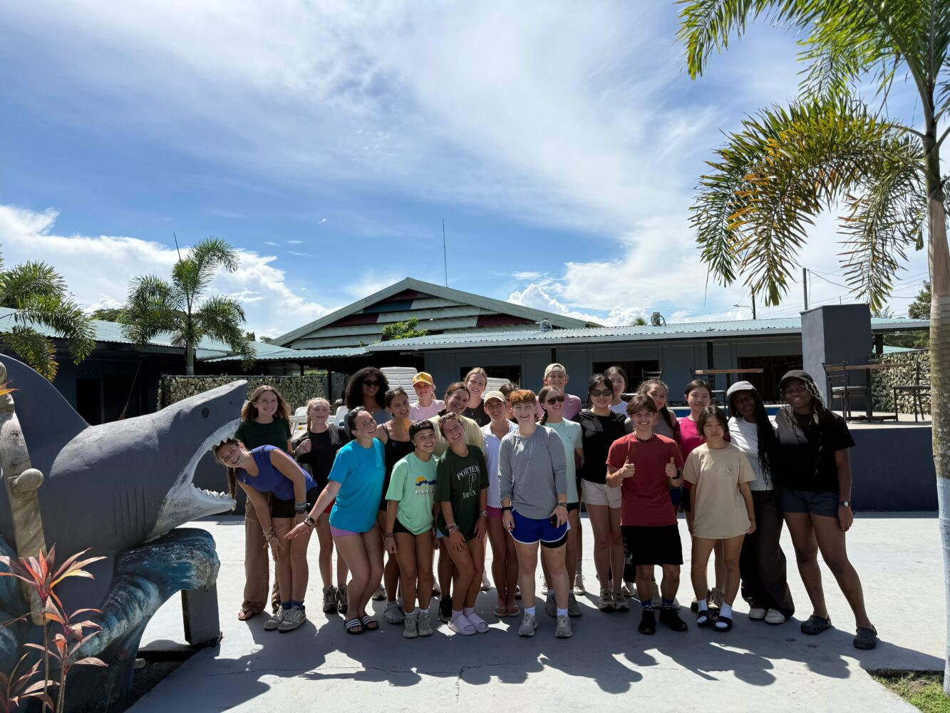 A large group of young people standing together outdoors in front of a shark statue near a pool.