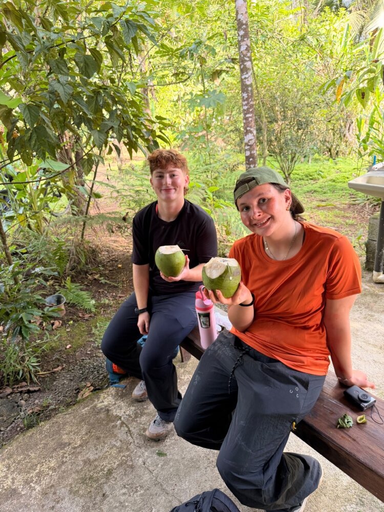 Two people sitting on a wooden bench in a wooded area, each holding a freshly opened green coconut.