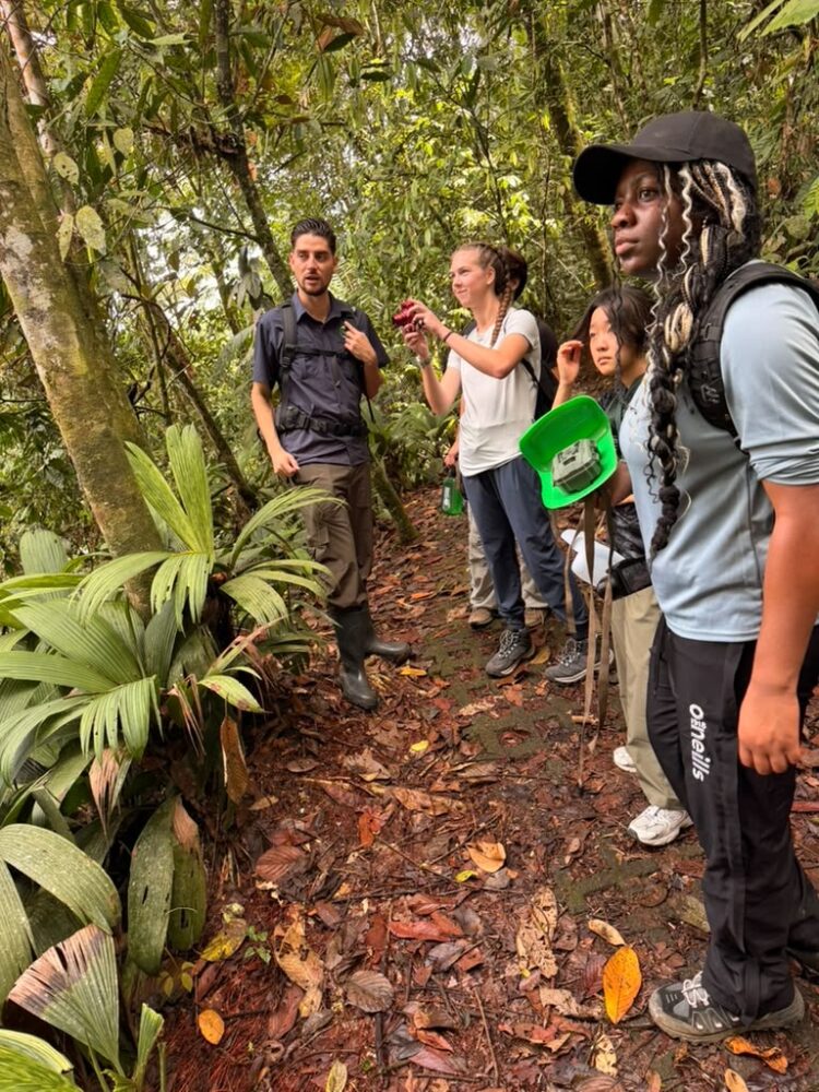 A guide stands on a forest trail with a group of four people, one of whom holds a green wildlife camera.