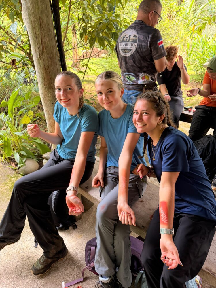 Three young women sitting on a bench, each displaying a red fern-shaped temporary tattoo on their forearm.