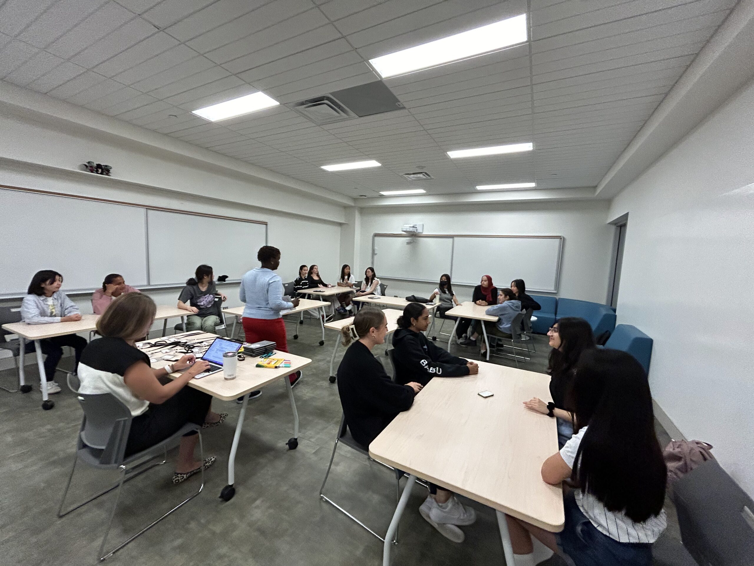 A classroom with students seated at desks in groups for Voices of Leaderhsip, listening to a teacher standing near a whiteboard. The room is bright with overhead lights and white walls.