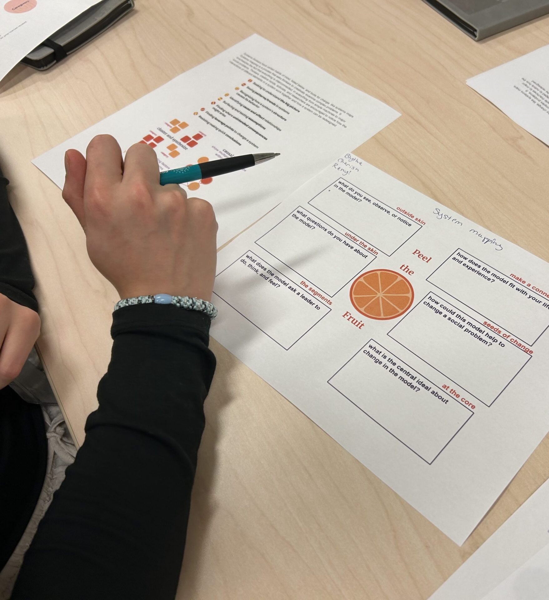 A person writes on a worksheet with diagrams and text about fruit while holding a pen. Several printed papers are spread out on the light wood desk.