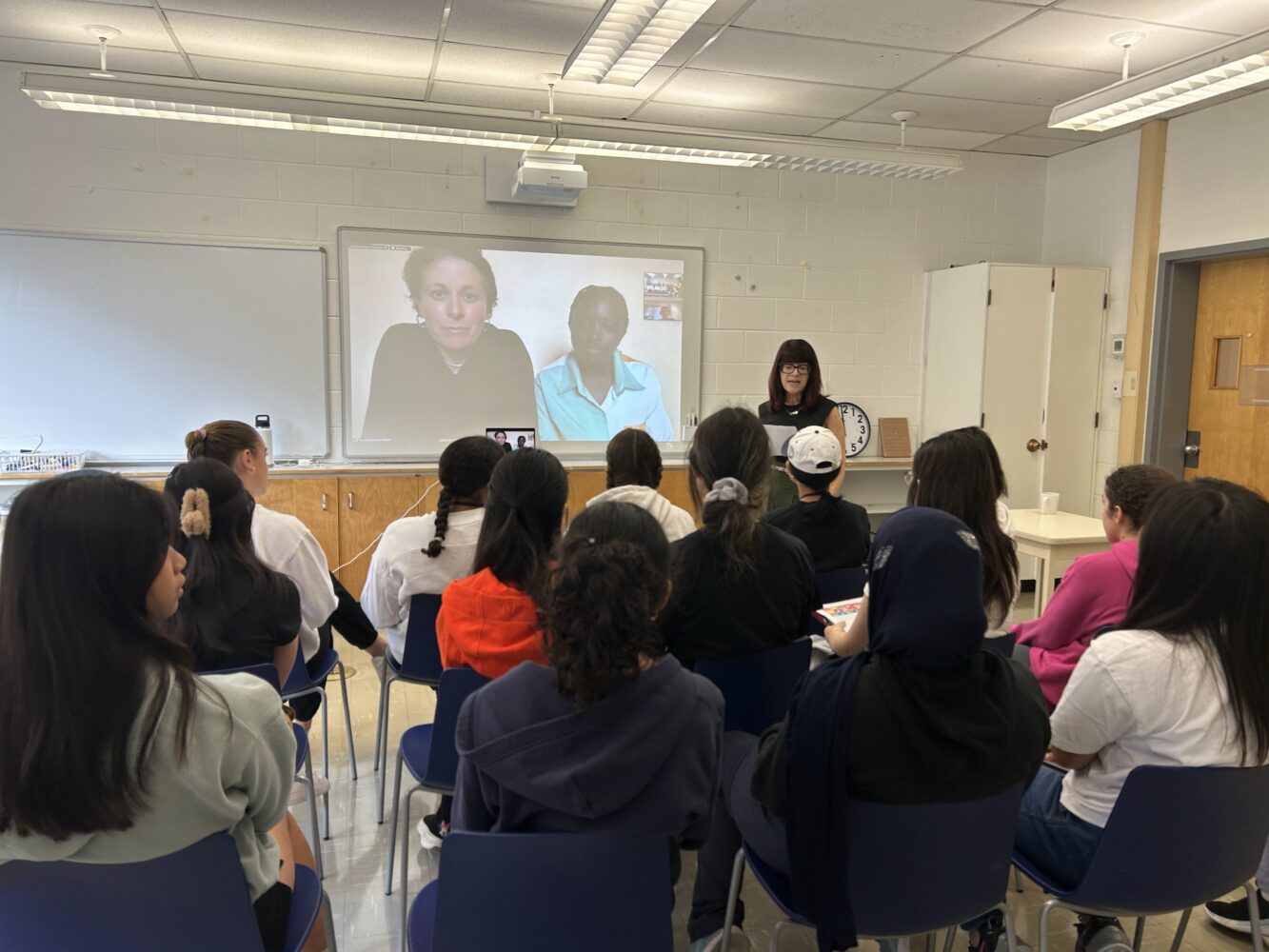 A classroom of students listens to a teacher speaking at the front, with two people appearing on a video call projected onto a whiteboard behind her.