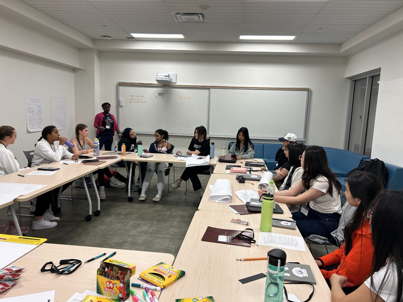 A group of students sits around tables in a classroom, engaged in discussion. Papers, water bottles, and art supplies are on the tables. One person stands near the whiteboard at the front of the room.