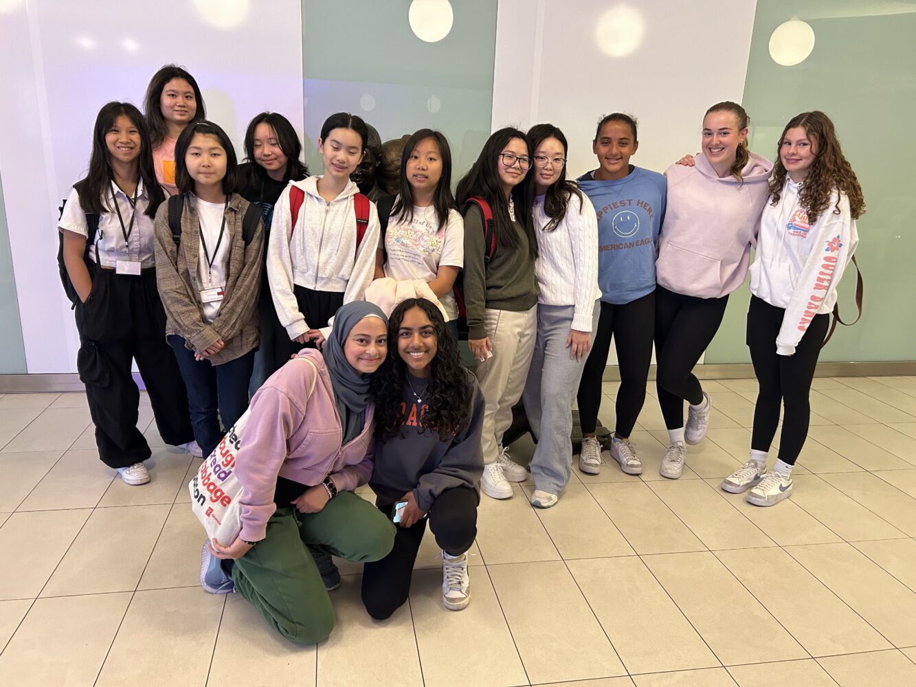 Thirteen teen girls stand together indoors, smiling for a group photo. Most are casually dressed, and some wear glasses or headscarves. The background has white walls and round decorative panels.