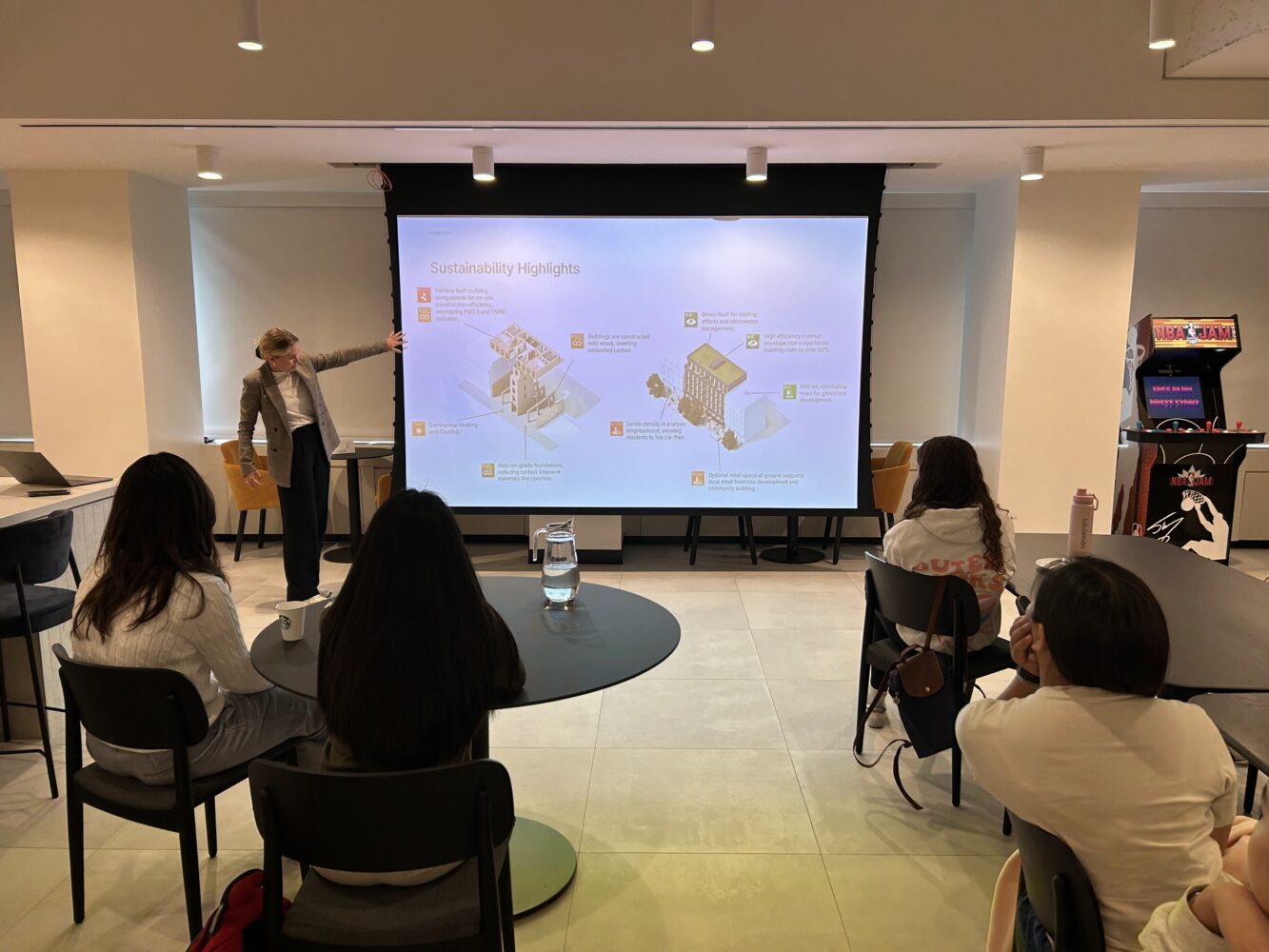 A woman presents sustainability highlights on a projector screen to a small group seated at tables in a modern meeting room. An arcade machine is visible in the background.
