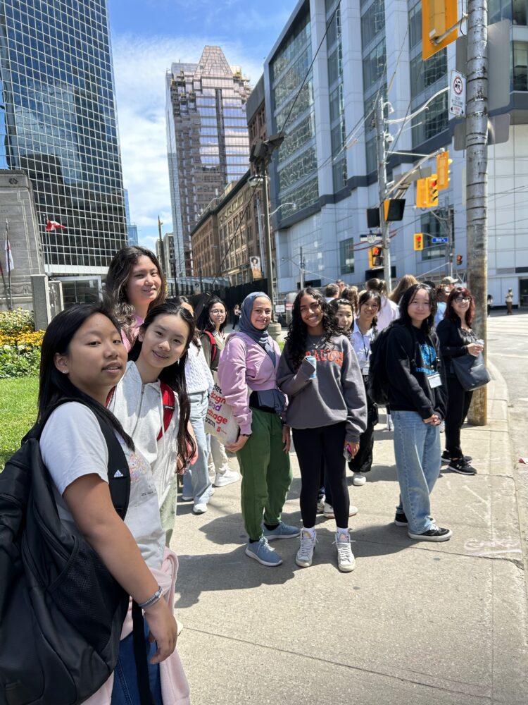 A group of young people stand together on a sunny city sidewalk, smiling at the camera. Tall buildings and traffic lights are visible in the background.