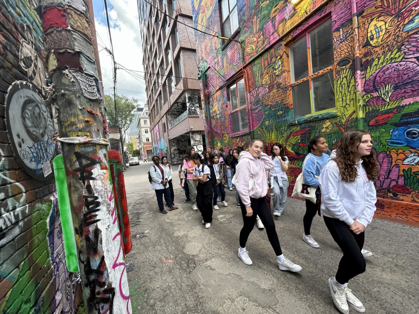 A group of teens walk through a colorful graffiti-filled alley, with bright murals covering the walls and urban street art all around them.