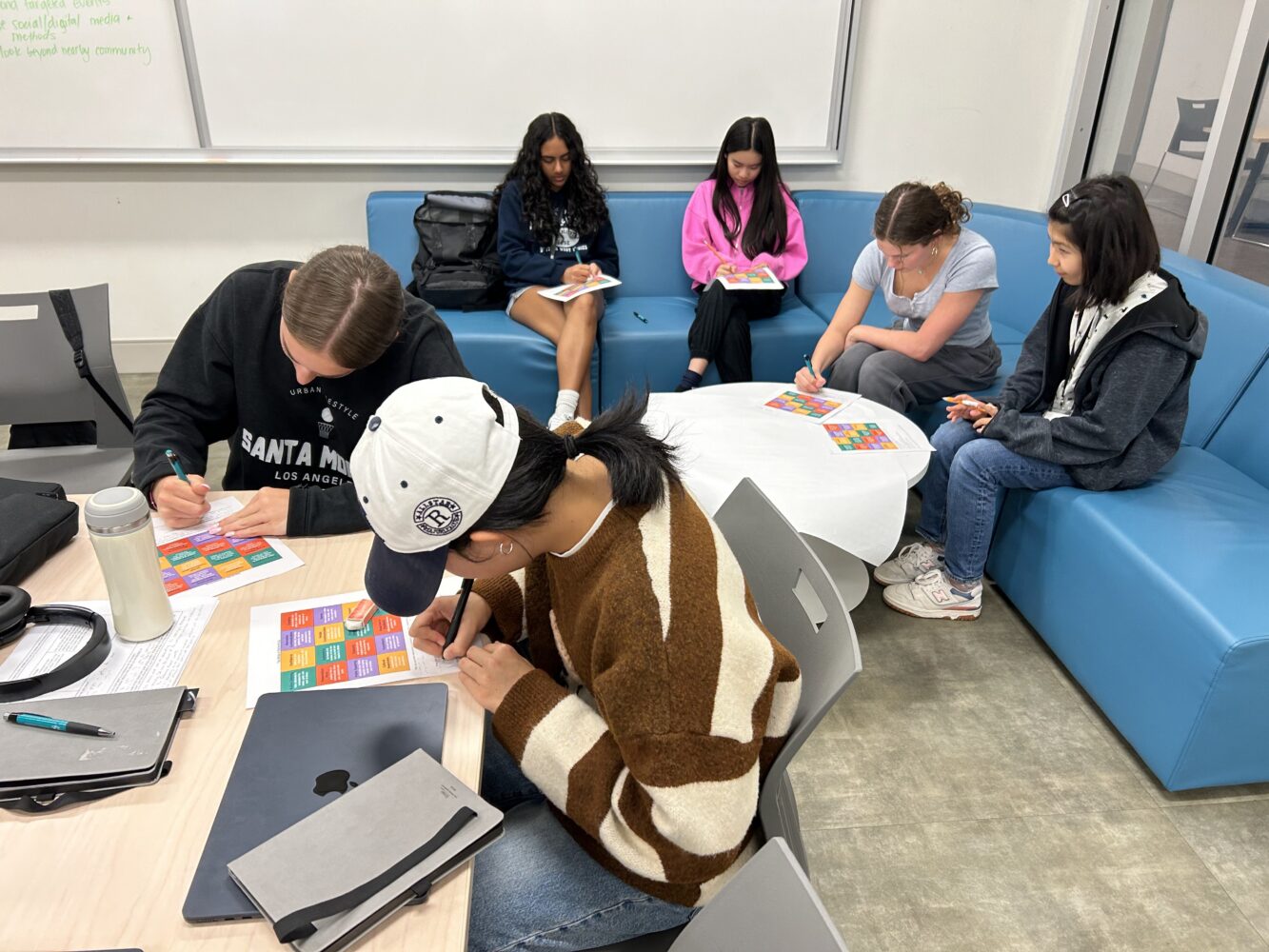 Six students sit around a table and on couches in a classroom, writing or working on colorful worksheets. Laptops, papers, and water bottles are also visible on the table.