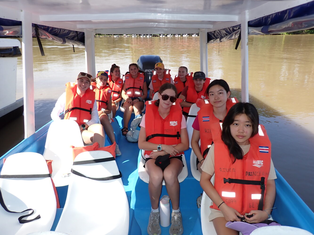 A group of people wearing orange life vests sit inside a boat on a river.