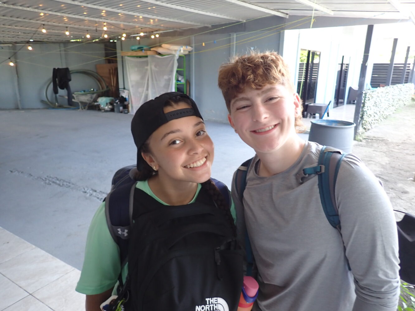 Two teenagers wearing backpacks smiling side by side for a photo.
