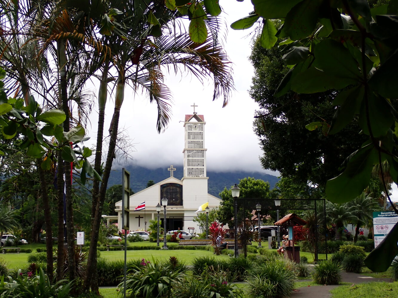 A white church with a tall bell tower and a cross, framed by palm trees and foliage, with a cloudy mountain in the background.