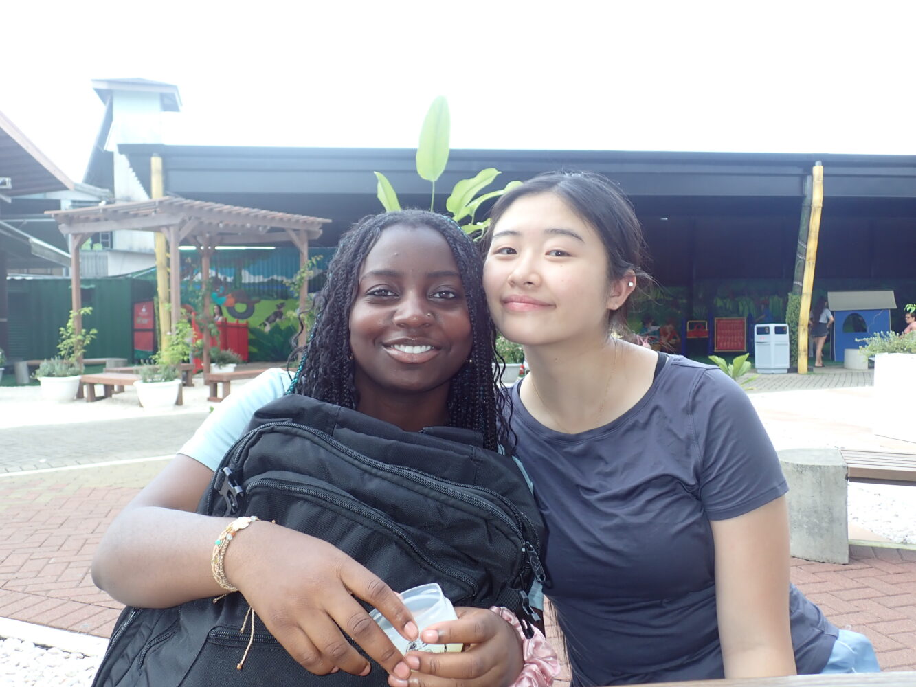 Two young women smiling and leaning toward each other while sitting outdoors.