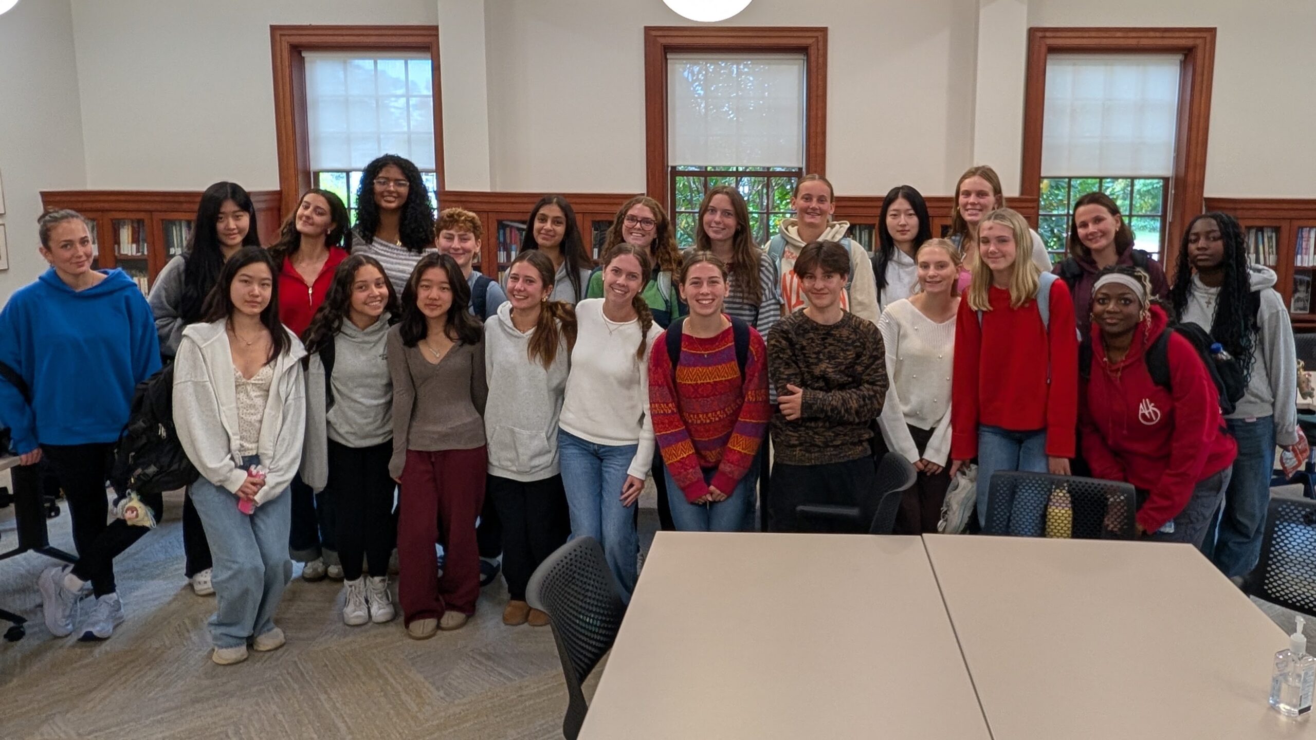 A group of twenty-five people, mostly young women, stand closely together and smile in a classroom with bookshelves, tables, and large windows.