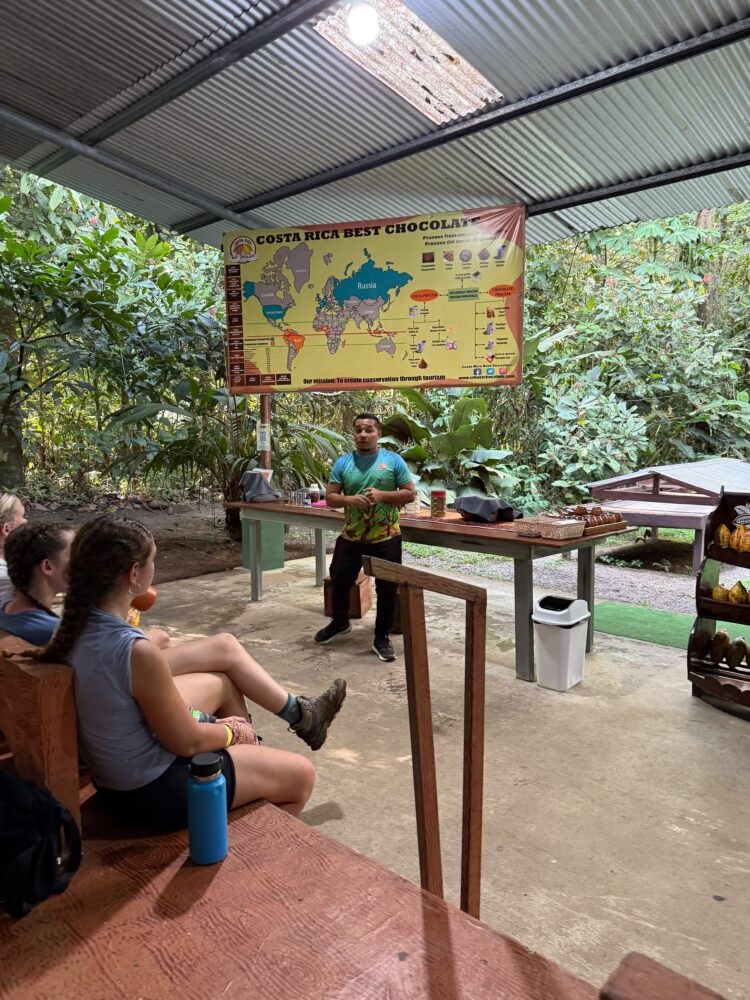 A man stands in an open-air pavilion giving a presentation in front of an educational poster about the chocolate-making process, while a small group of people sits on benches listening.