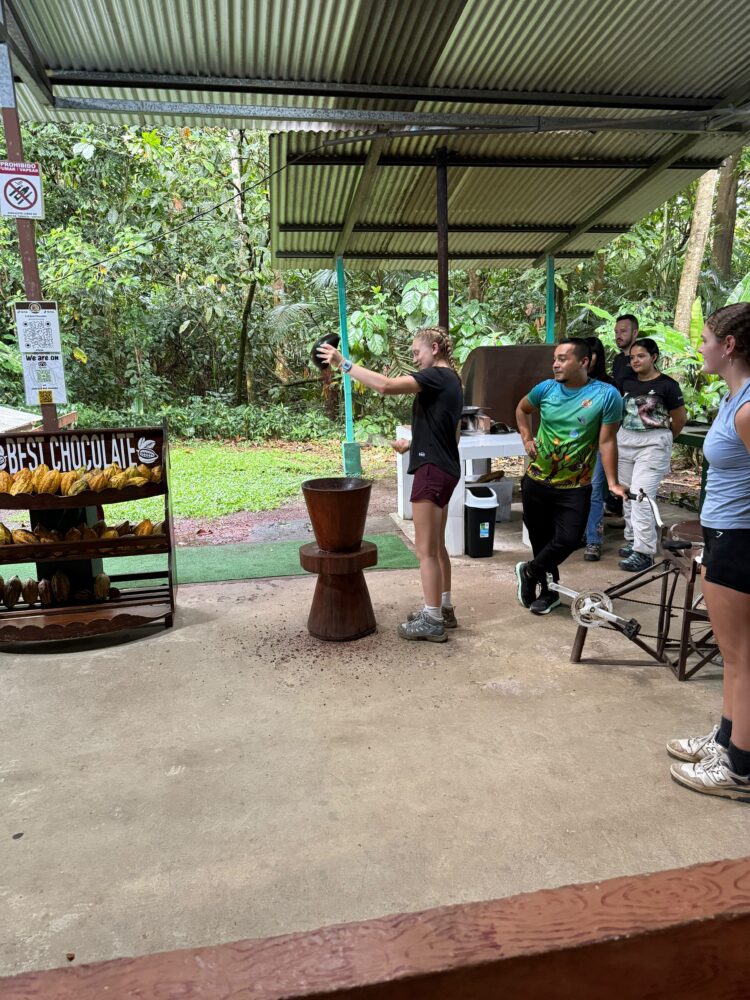 A woman demonstrates using a wooden mortar and pestle to a small group in an outdoor cocoa processing area with shelves of cacao pods nearby.