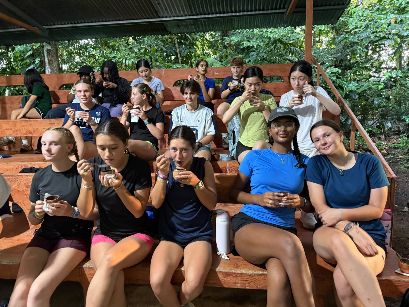 A group of young people sitting on wooden bleachers outdoors, drinking from small cups.