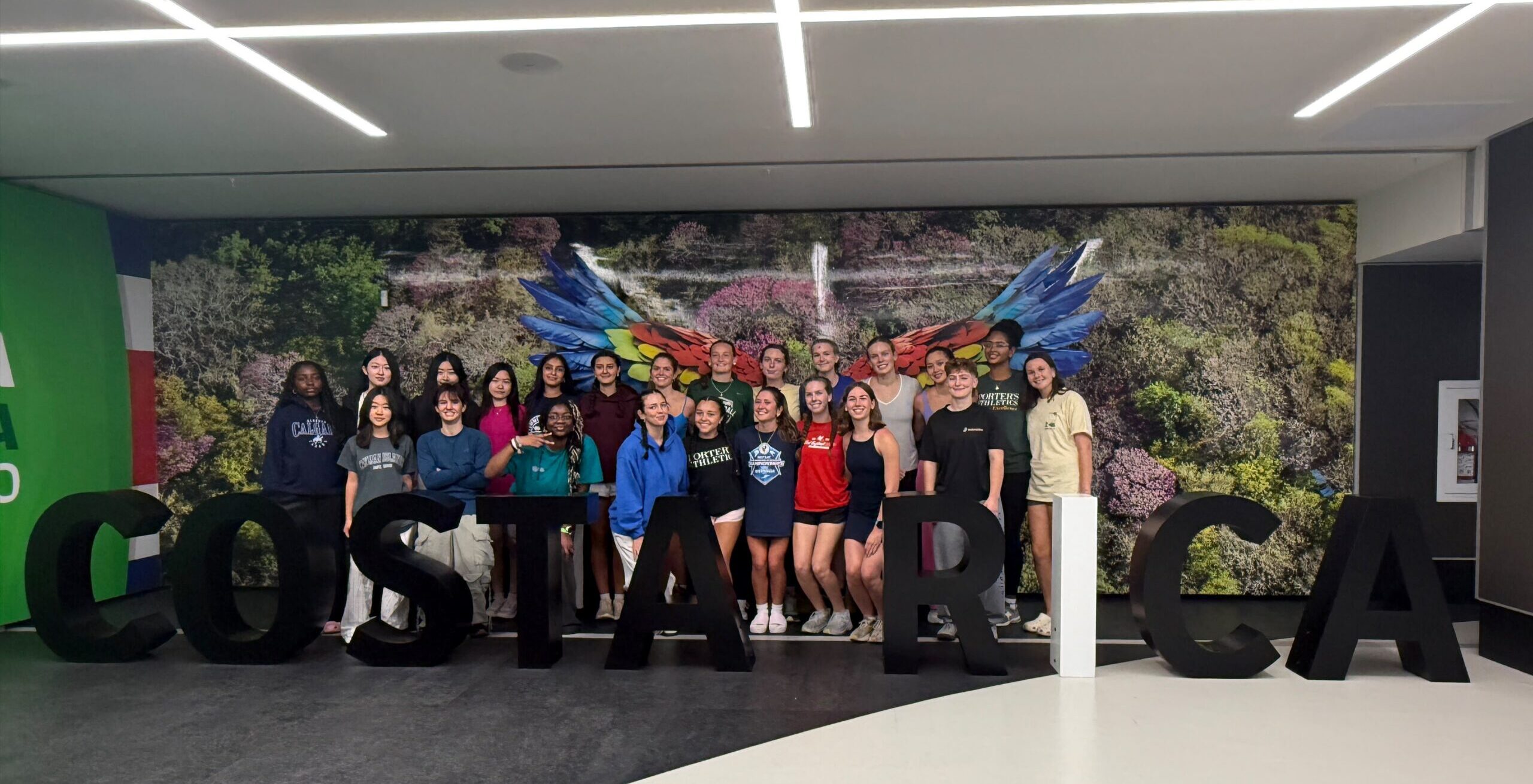 A large group of students posing in front of a giant COSTARICA sign with a mural of macaw wings in the background.