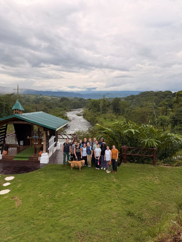 A group of fourteen people and a golden dog pose for a photo on a grassy lawn next to a small wooden chapel overlooking a river and forest.
