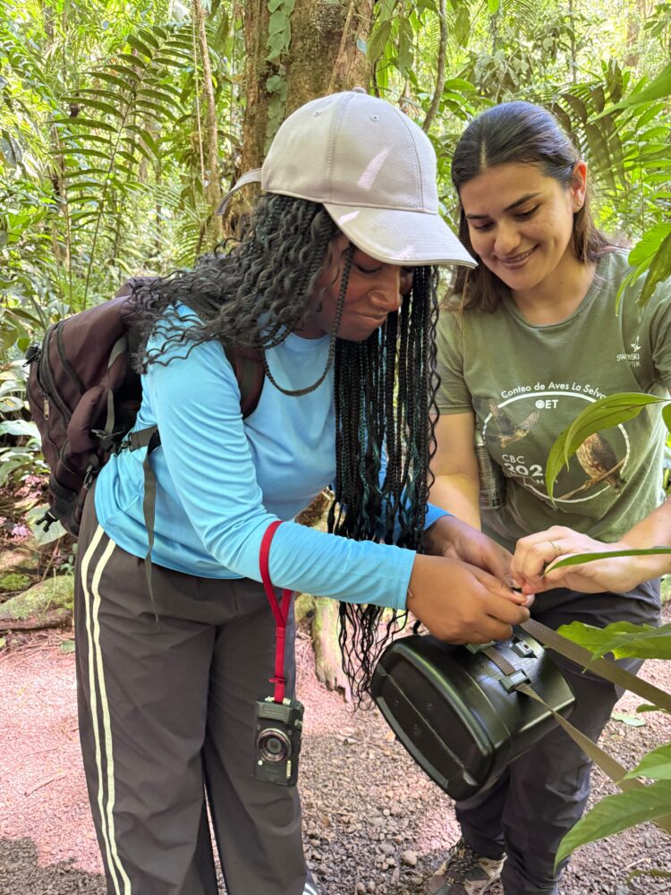 Two women in a forest working together to adjust a camera device mounted on a tripod.
