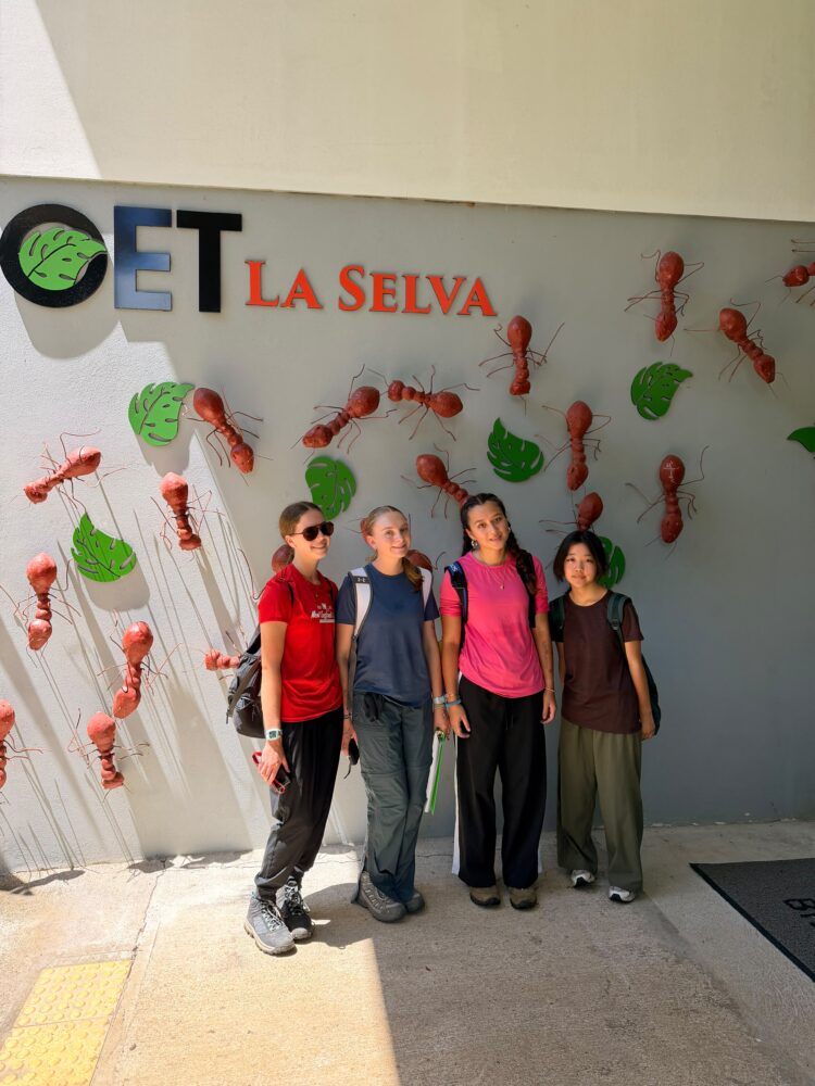 Four young people stand in a row in front of a wall decorated with metal ants and green leaves, below a sign reading La Selva.