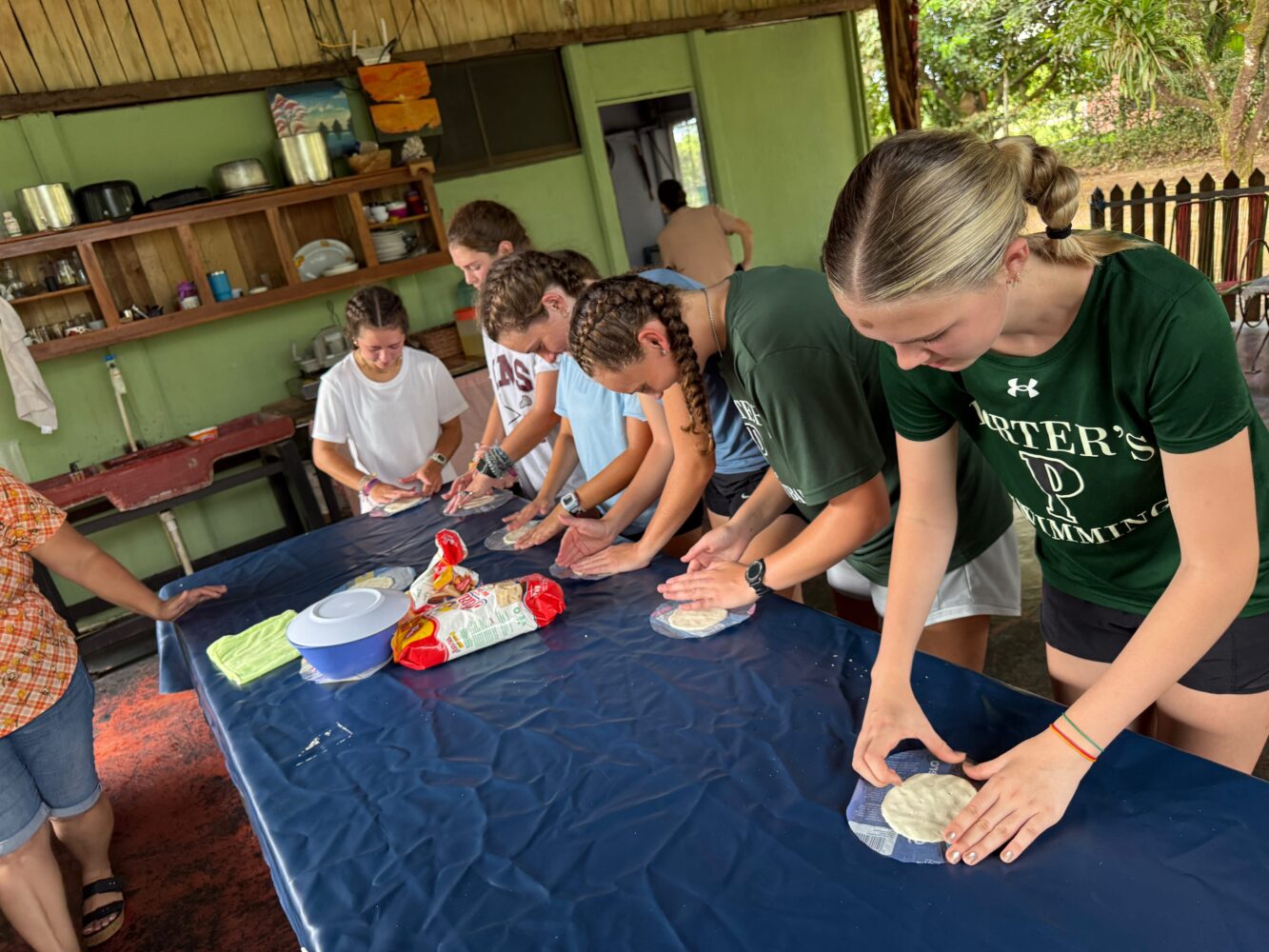 Five young women stand in a row at a blue-covered table, flattening dough onto small squares of plastic.