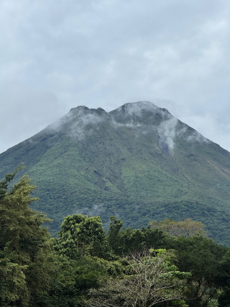A tall, green, cone-shaped volcano partially covered by clouds, rising above a forest canopy.