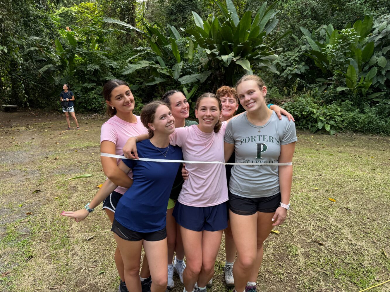 Six young women stand together with their arms around each other behind a white finish line ribbon in a forested area.