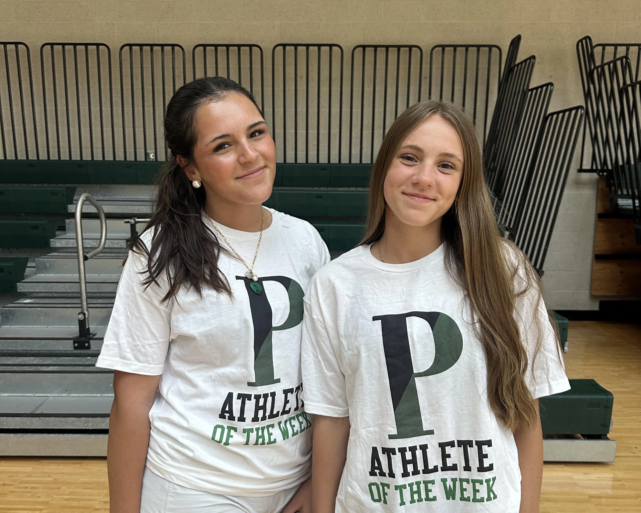 Two young women wearing white T-shirts that say P Athlete of the Week stand in front of gymnasium bleachers.